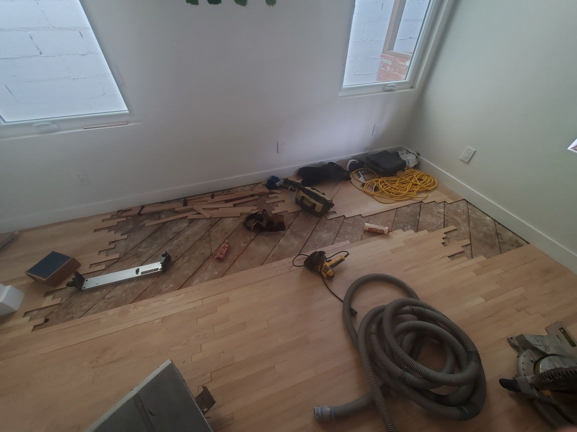 A room with light wood flooring, featuring a partially removed section of planks and scattered construction tools.