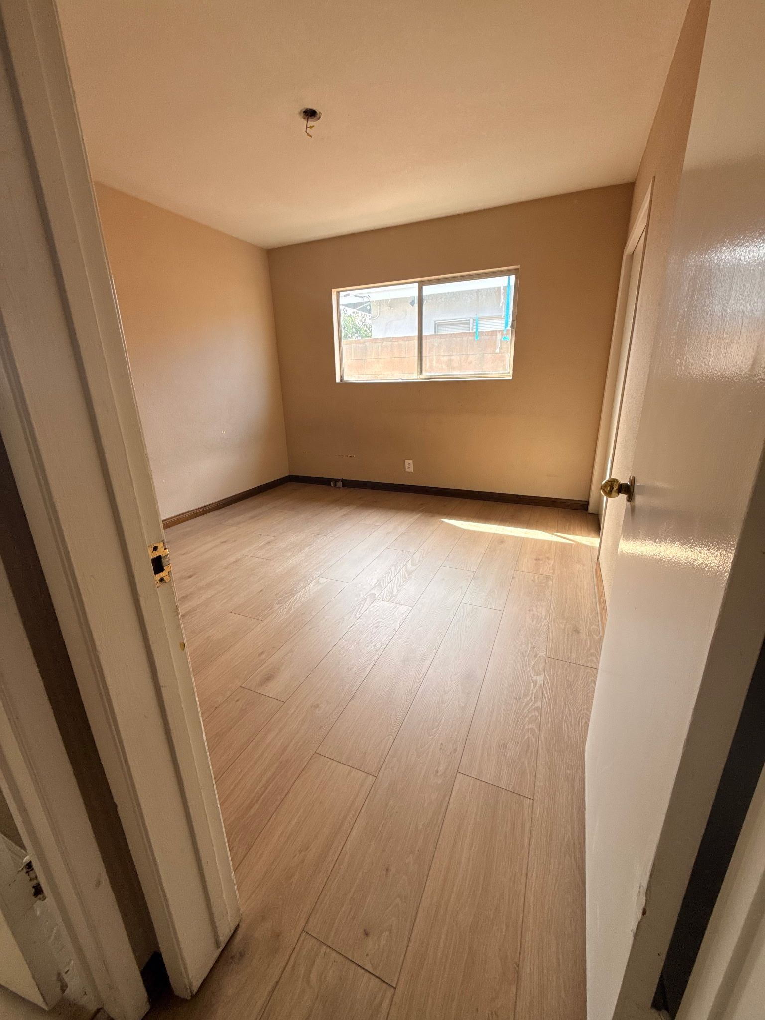 An empty bedroom with light-colored laminate flooring, beige walls, and a single window looking out onto a fence.