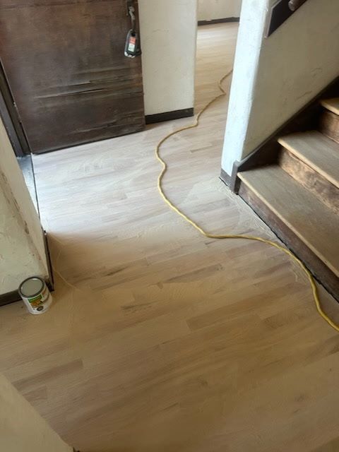 Freshly sanded, light-colored hardwood floor in an entryway with a wooden door, stairs, and a yellow extension cord.