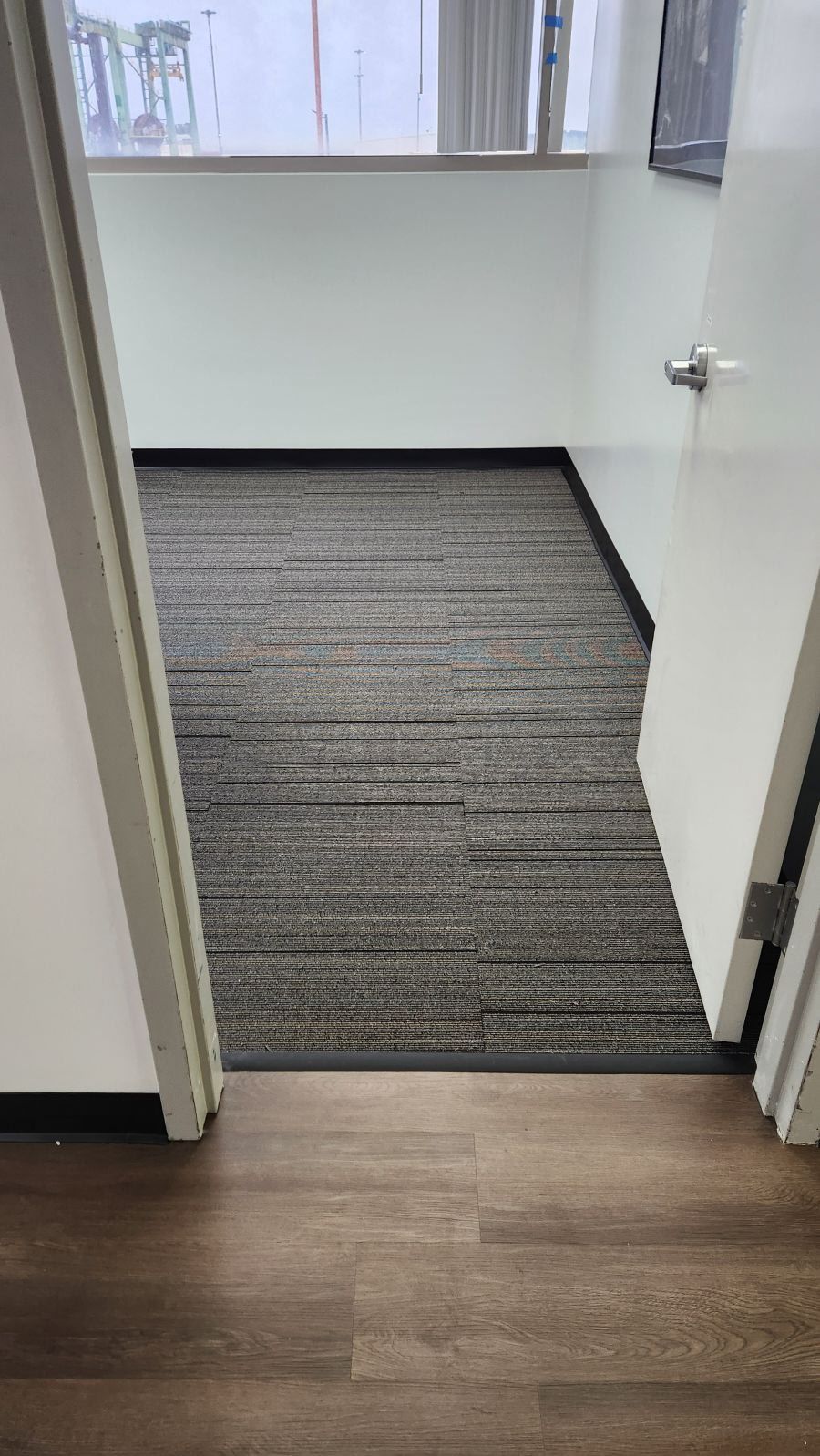 A doorway view into a small office space with dark grey carpet, white walls, and wood-look flooring in the foreground.