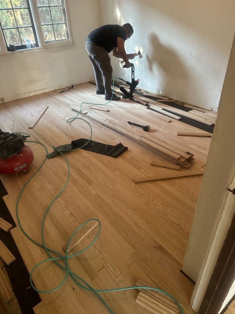 A worker installs hardwood flooring in a room, using a pneumatic tool to nail planks against the wall.