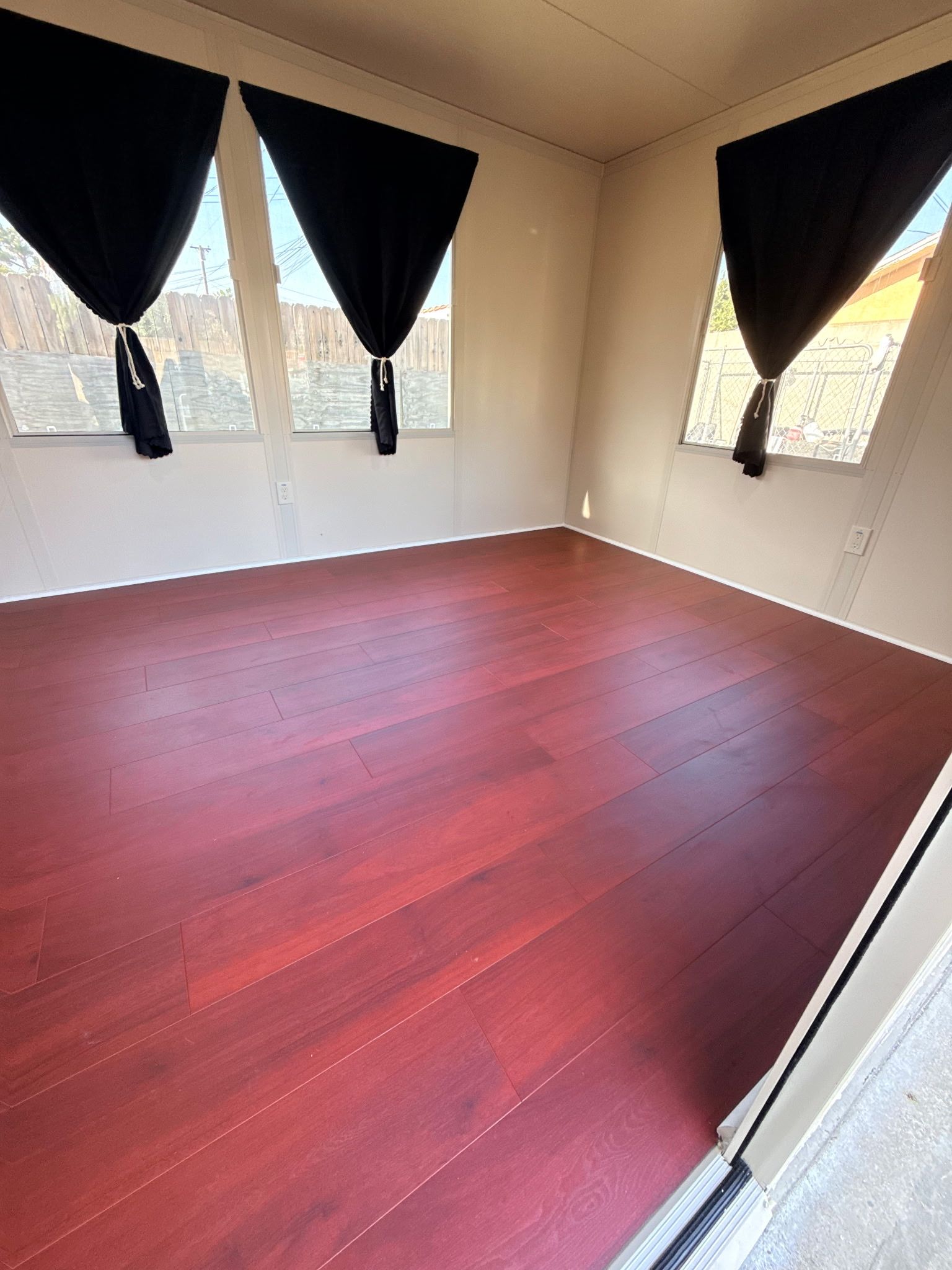 An empty room with dark red laminate floors, light-colored walls, and black curtains on three windows.
