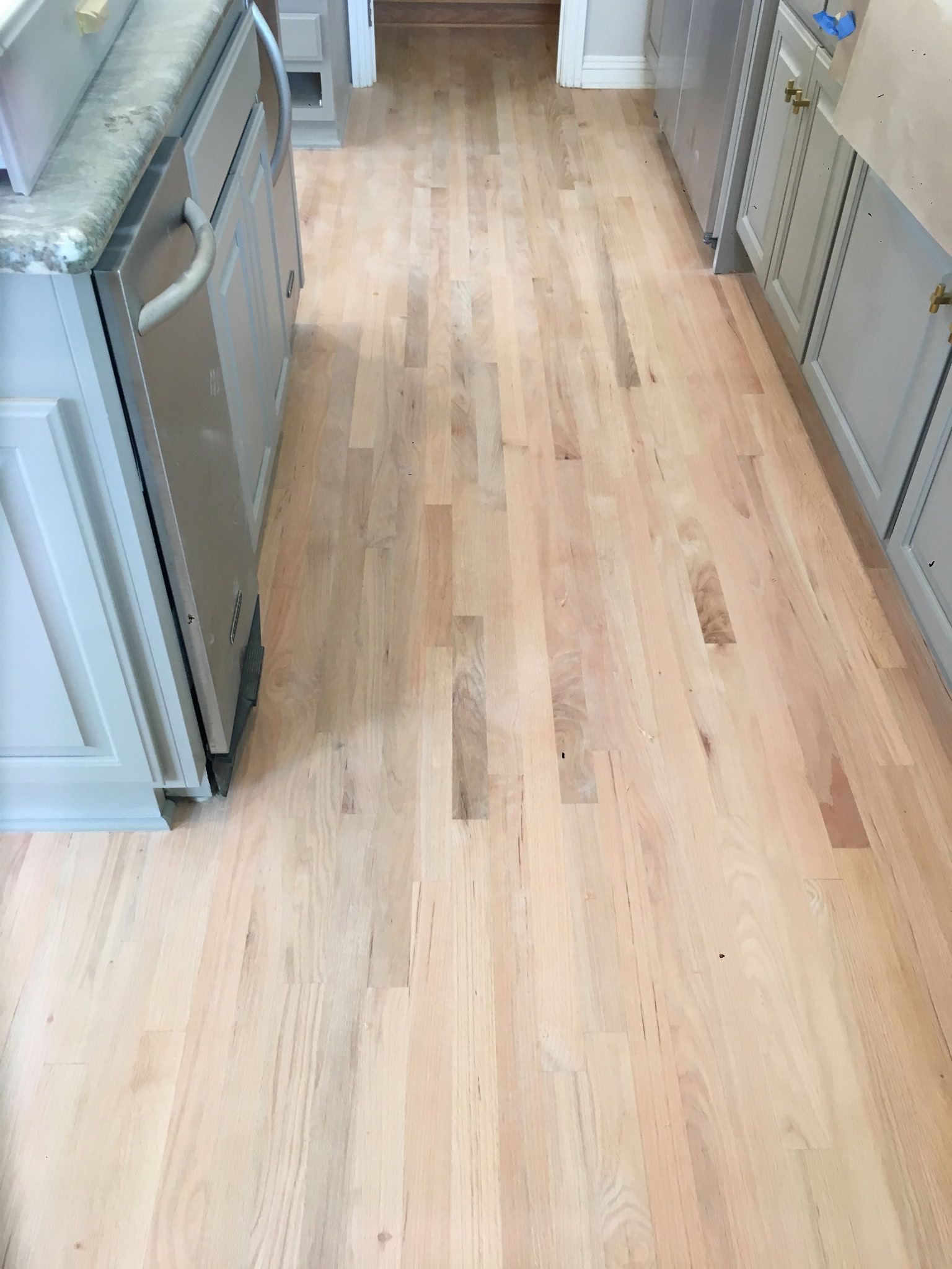 A narrow kitchen walkway with light-toned hardwood flooring installed between two rows of grey kitchen cabinets.