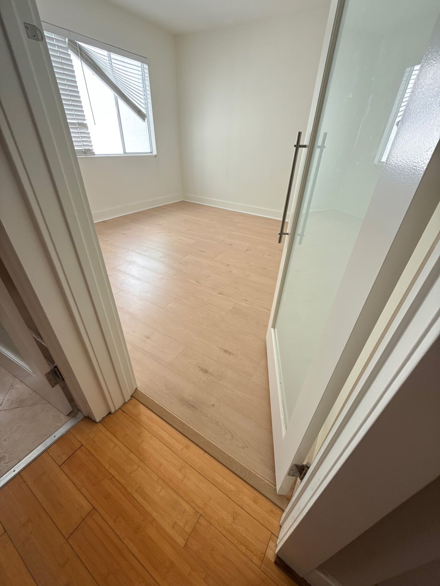 A view from a doorway into an empty room with light-toned wood flooring, white walls, and a window with white blinds.