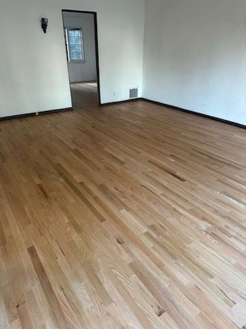 A bright, empty room with light-colored hardwood floors, white walls, and a doorway leading to a hallway.