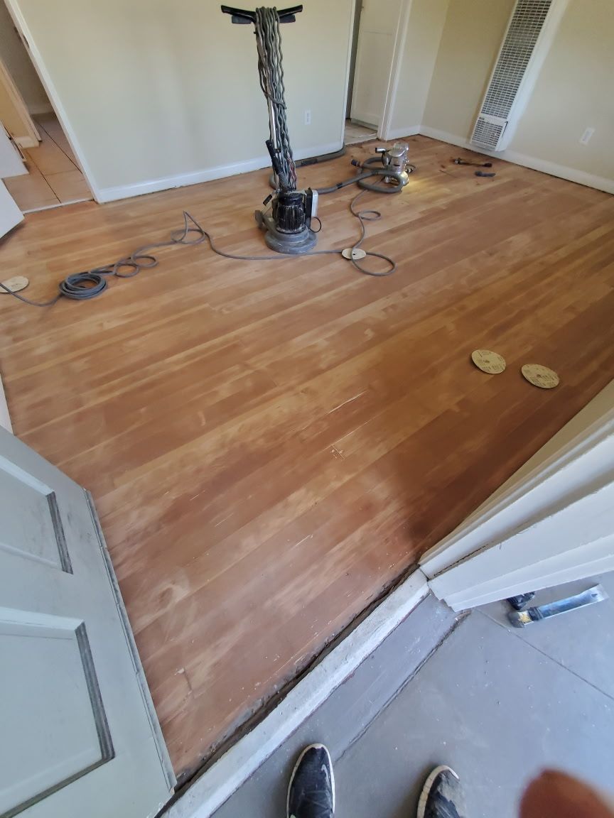 A room with wooden flooring being sanded, featuring a floor buffer machine, cords, and sanding discs on the unfinished wood.