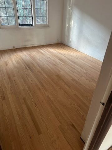 A bright, empty room featuring newly refinished light-toned hardwood floors and white walls with a large grid window.