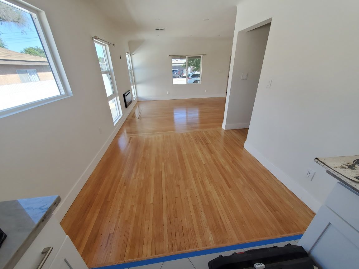 An empty room with polished light-wood floors, white walls, and several windows, viewed from a kitchen doorway.