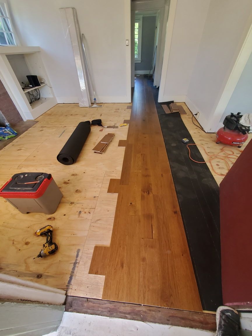 Half-installed wooden flooring in a room under renovation, with tools and underlayment visible on the bare plywood subfloor.