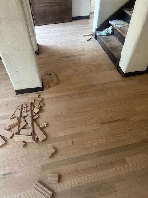 Hardwood flooring in a hallway with scrap wood pieces scattered on the floor near a pillar and a staircase.
