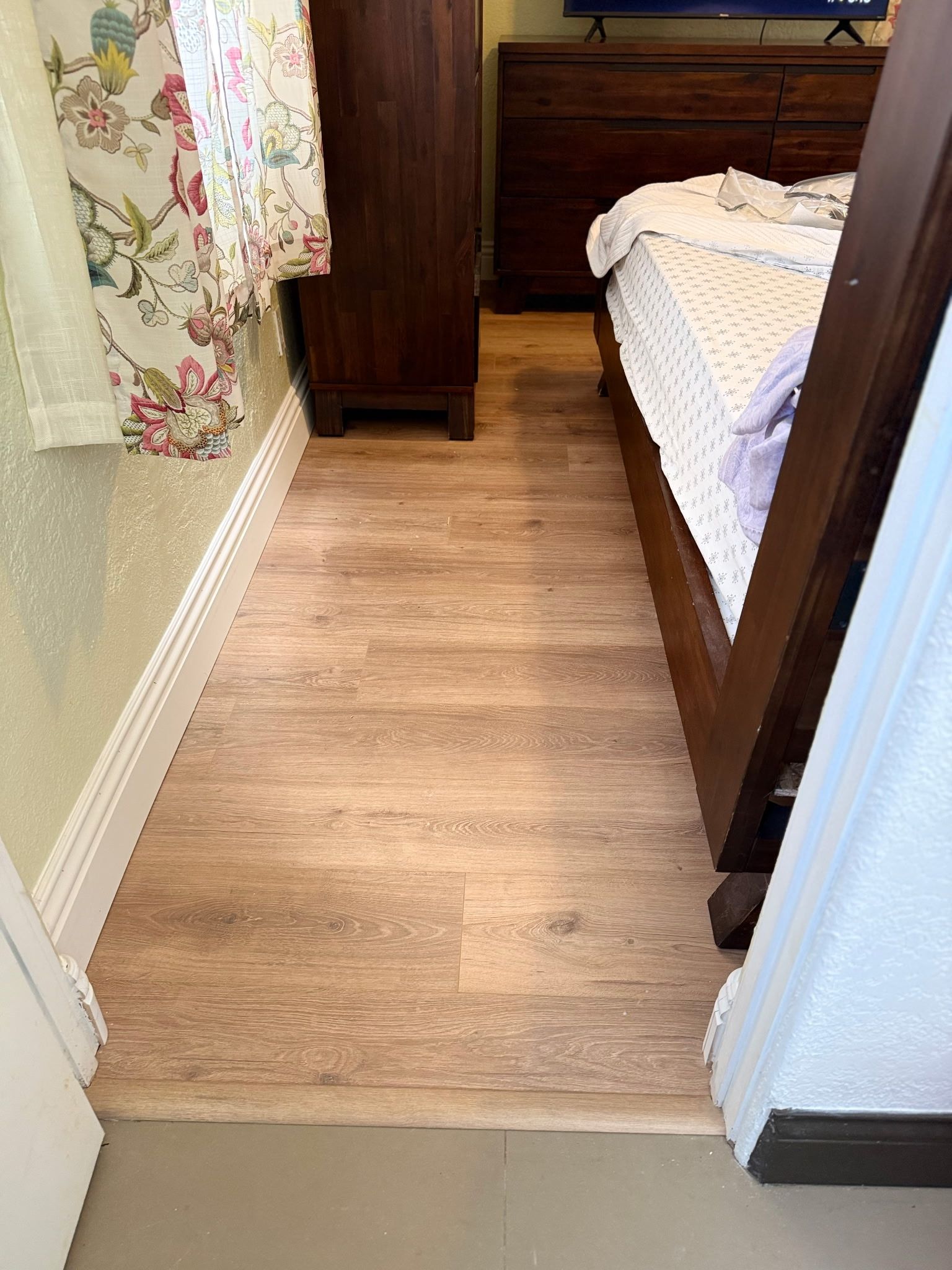 A hallway view of a bedroom with light wood-look flooring, a floral curtain on the left, and a wooden bed frame on the right.
