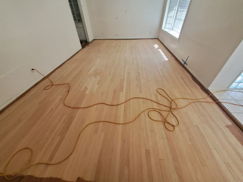 A room with newly sanded, unfinished light-wood flooring and a long orange extension cord running across it.