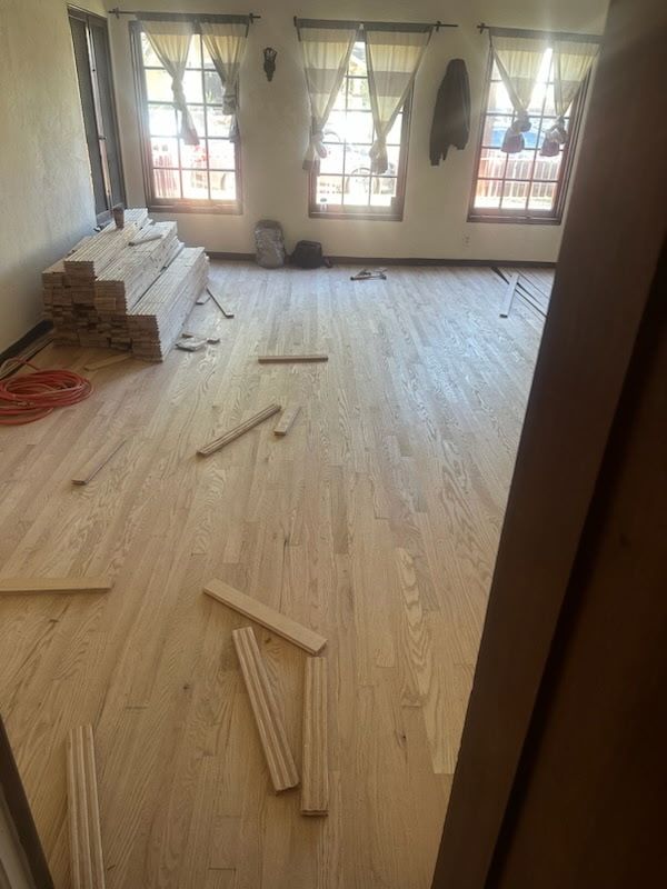A room under renovation with newly installed light-colored hardwood flooring, scattered wood scraps, and stacked planks.