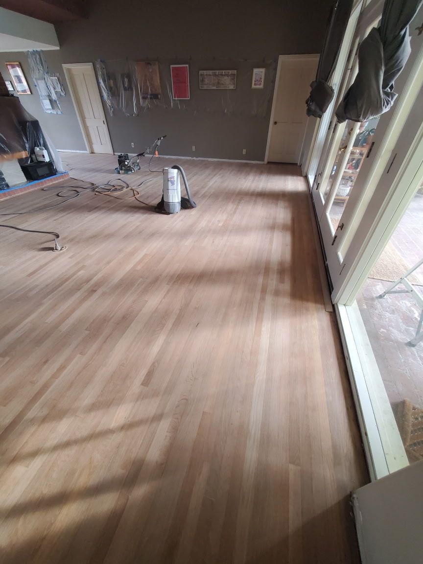 A light-colored wood floor in a room under renovation, featuring a floor sander in the center and large windows.