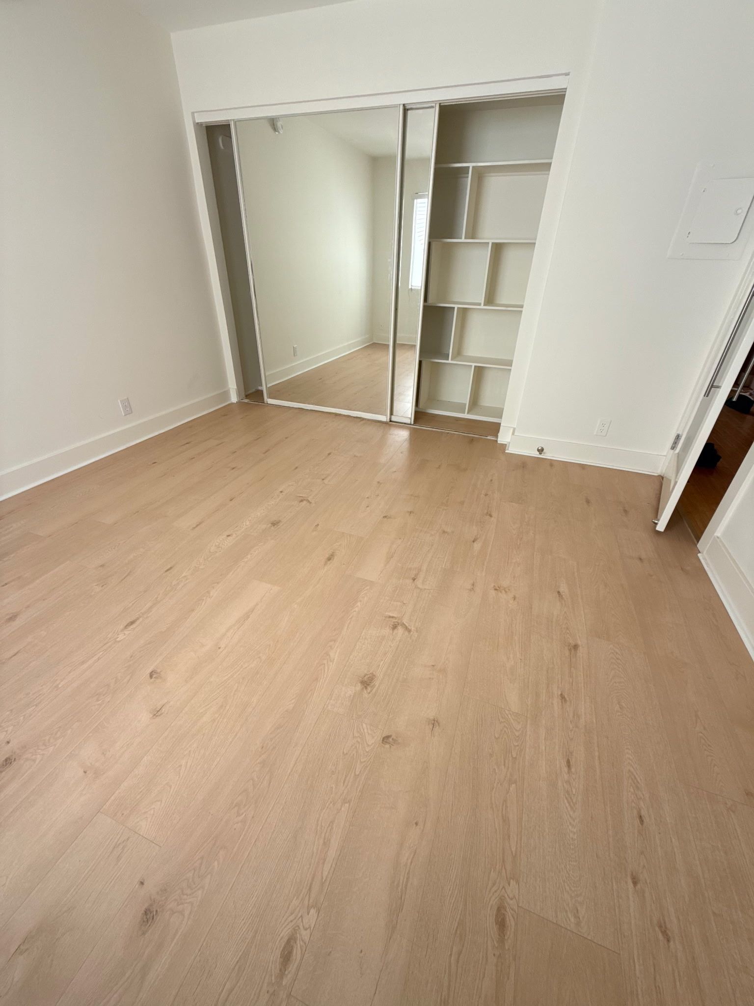 A light wood-floored room with white walls, a mirrored sliding closet, and a white built-in shelving unit.