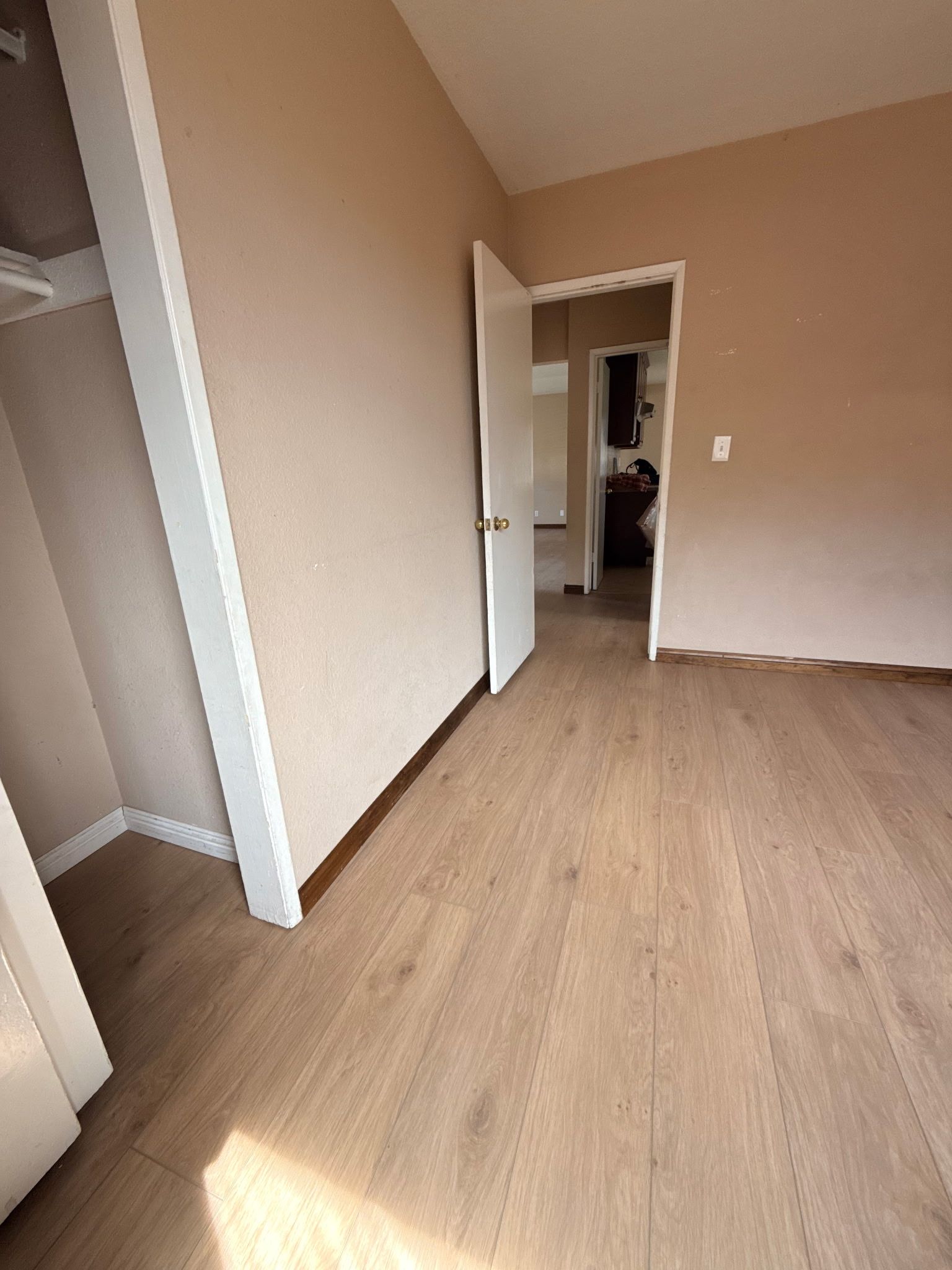 An empty room with light wood-look flooring, beige walls, and an open doorway leading to another room.