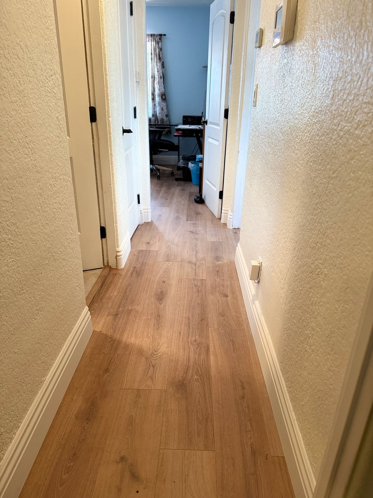 A hallway with light-colored, textured walls and wood-patterned flooring, leading toward an open doorway into a room.