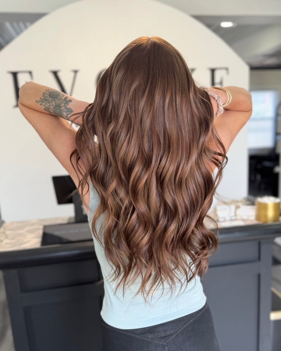 Woman with long, wavy brown hair, arms raised behind her head. Wearing a light blue top in a salon.