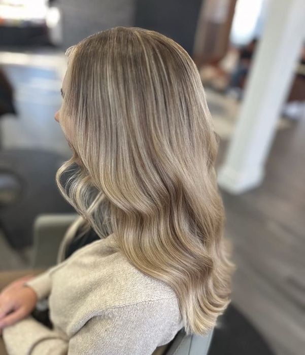 Woman with long, wavy blonde hair styled with highlights, sitting in a salon setting. She is wearing a beige sweater.