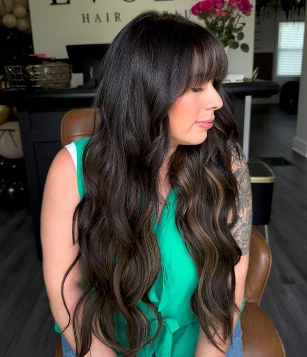 Woman with long, wavy dark brown hair and bangs, wearing a green top, sitting indoors.