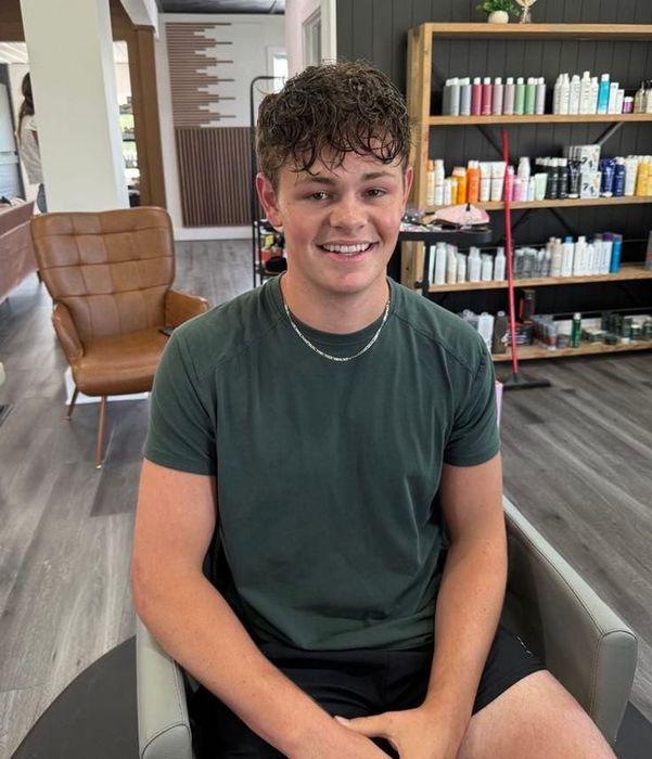 A young man with curly brown hair smiles at the camera, seated in a salon chair. He wears a green t-shirt and silver necklace.