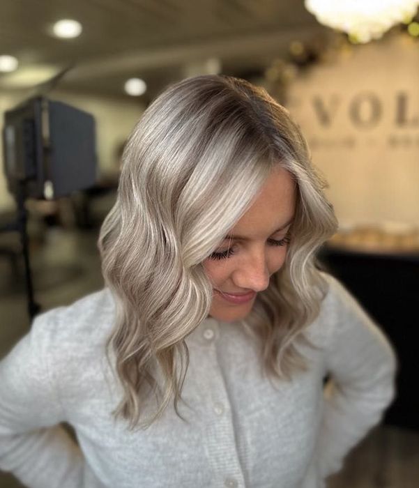 Woman with wavy, blonde hair smiles, wearing a cream sweater, in a salon setting.