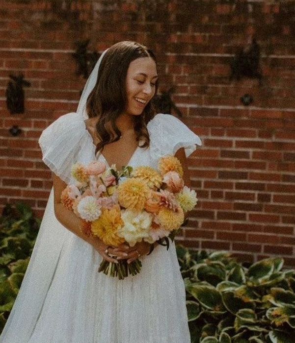 Bride smiling, holding a colorful bouquet, stands in front of a brick wall, wearing a white dress and veil.