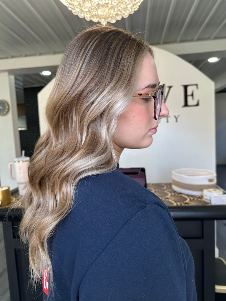 Woman with wavy, blonde hair and glasses, side view, in a salon with a dark blue shirt.