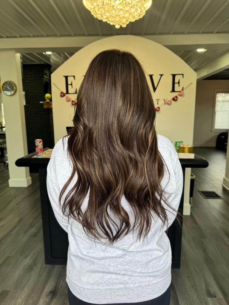 Woman with long wavy brown hair in a salon, standing in front of a black table, back view.
