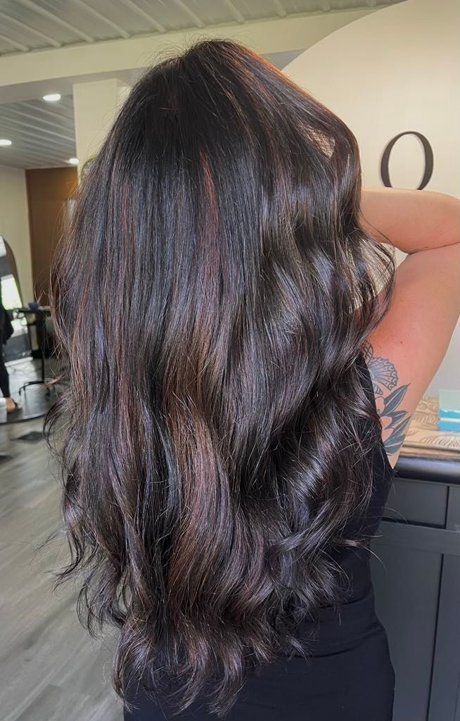 Woman with long, wavy, dark hair with highlights in a salon, reaching her arms up.