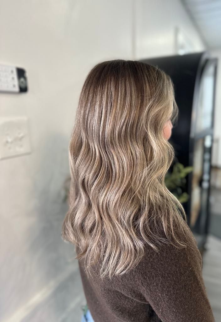 Woman with wavy, highlighted hair, brown and blonde, in a salon setting. She is wearing a brown sweater.