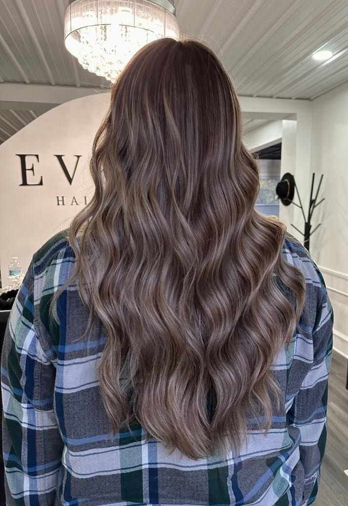 Woman with wavy, brown hair with highlights. She wears a plaid shirt, in a salon setting.