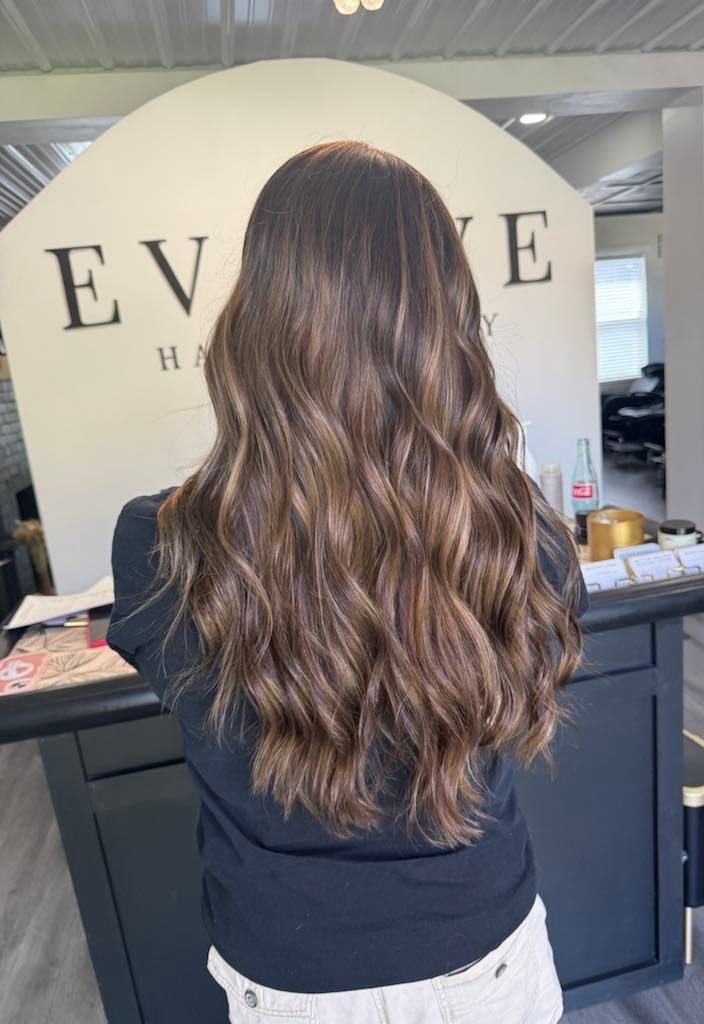 Woman with long, wavy, highlighted brown hair stands in front of a salon counter. Salon name 