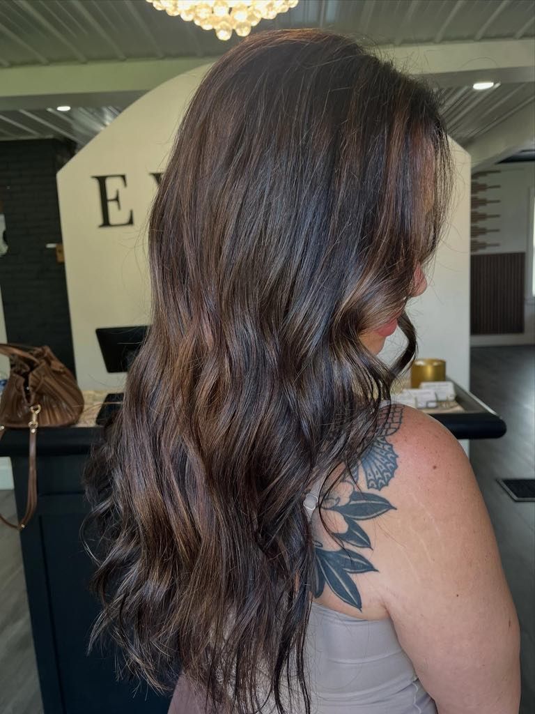 Woman with long, wavy brown hair, tattoo on shoulder, standing in a salon.