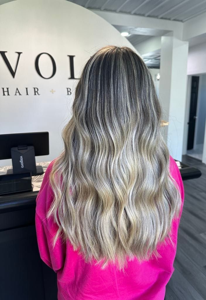 Woman with long wavy hair styled with balayage in a salon. Her hair is a mix of dark and light tones, wearing a pink sweater.