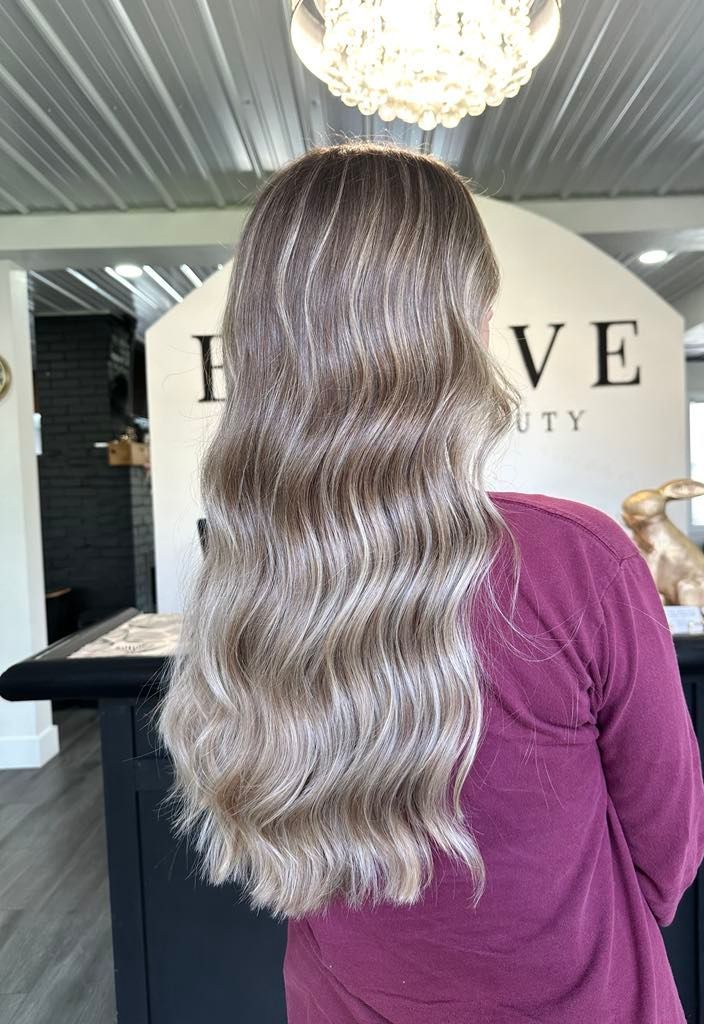 Woman with long, wavy blonde hair in a salon. She wears a maroon shirt and is standing in front of a counter.