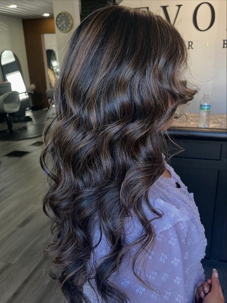 Woman with long, wavy, dark brown hair, with highlights, at a salon.