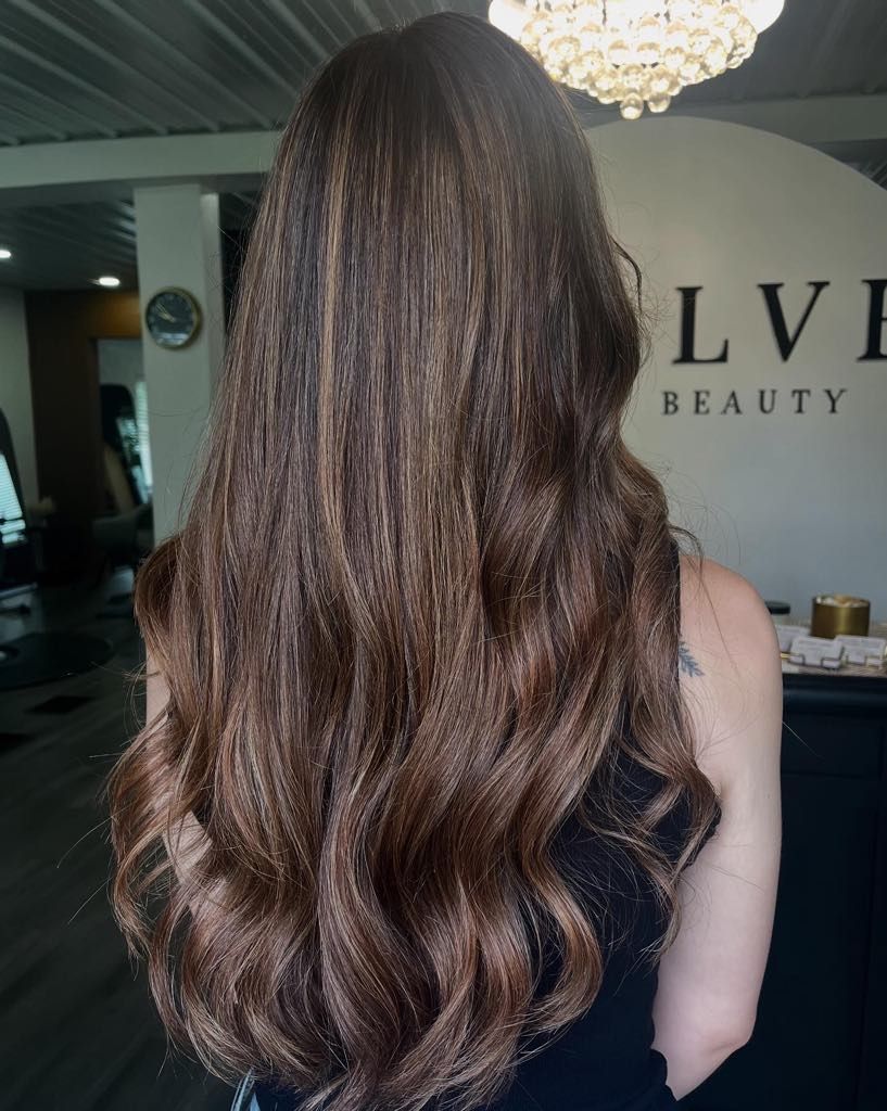 Woman with long, wavy, brown hair. Highlights, wearing black top, in a salon.