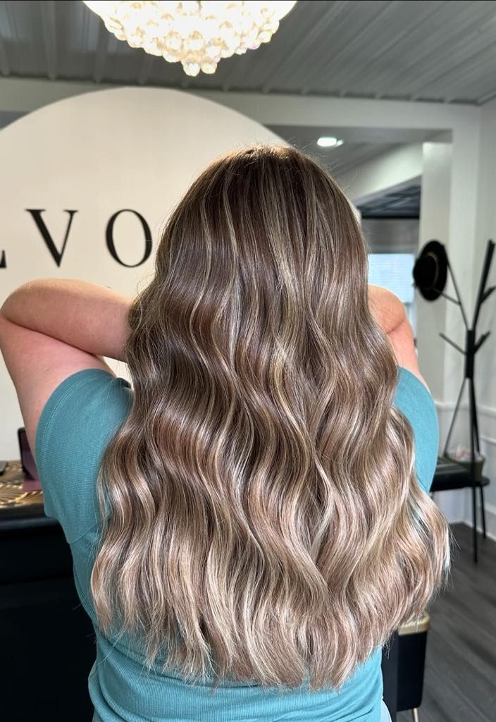 Woman with wavy, highlighted brown and blonde hair in a salon. She wears a teal shirt.