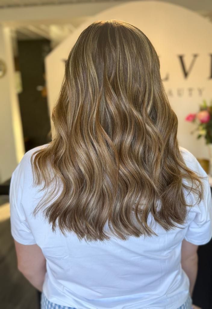Woman with wavy brown hair with blonde highlights; wearing a white t-shirt. In a salon setting.