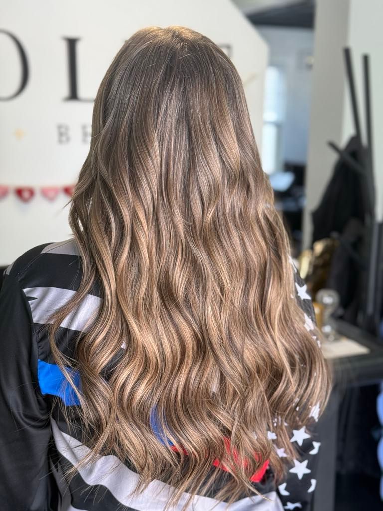 Woman with long wavy brown hair with lighter highlights in a salon. She wears a black and white striped top.