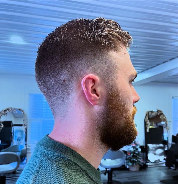 Man with a fade haircut and beard, profile view. Wearing a green sweater, inside a salon.