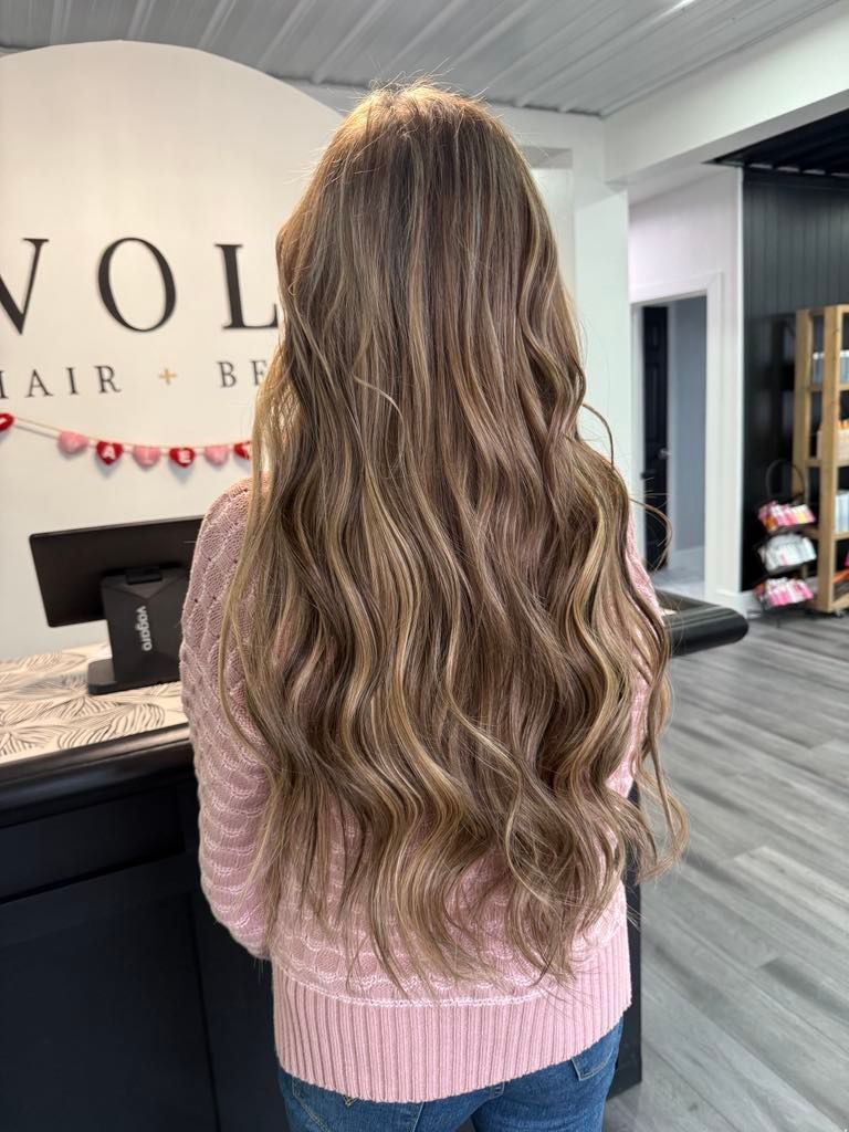 Woman with long, wavy brown hair in a salon, wearing a pink sweater and jeans. Back view.