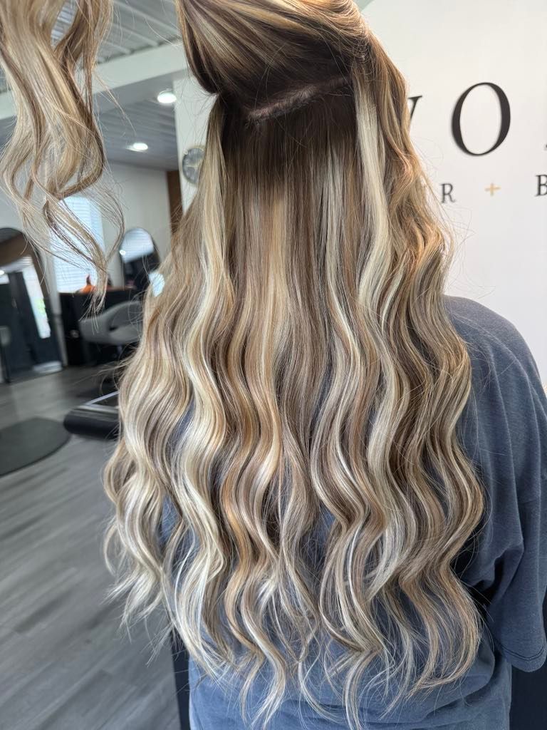 Woman with long wavy blonde and brown highlighted hair, standing in a salon.