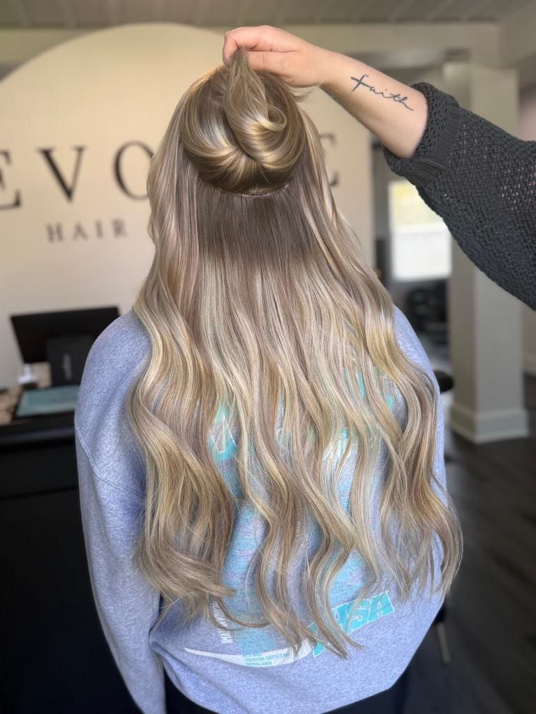 Woman with long blonde hair, styled with a half-up bun, stands in a salon. A hand with a tattoo reaches up to her hair.