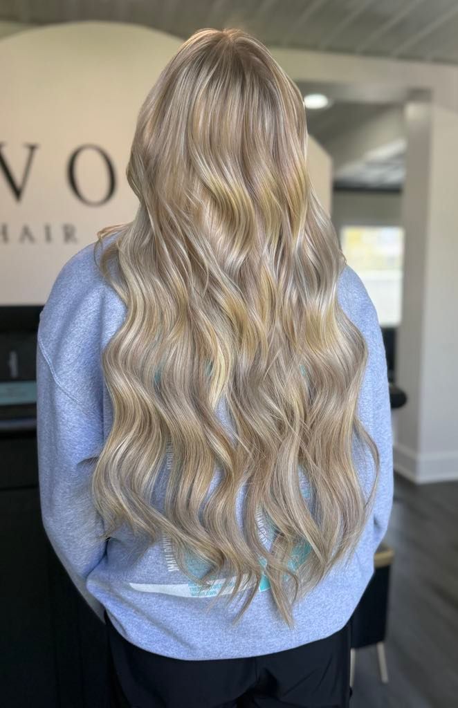 A woman with long, wavy blonde hair, wearing a blue sweatshirt, stands indoors with a white wall in the background.