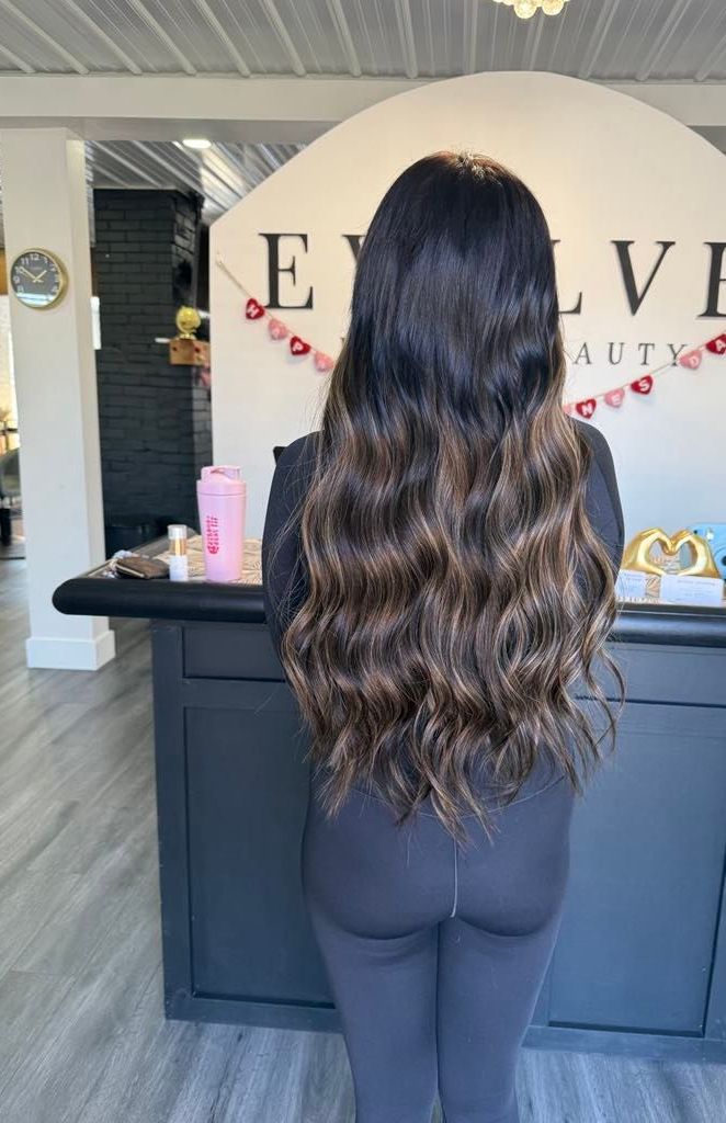 Woman with long, wavy dark hair stands near a black counter in a beauty salon. The salon's logo reads 