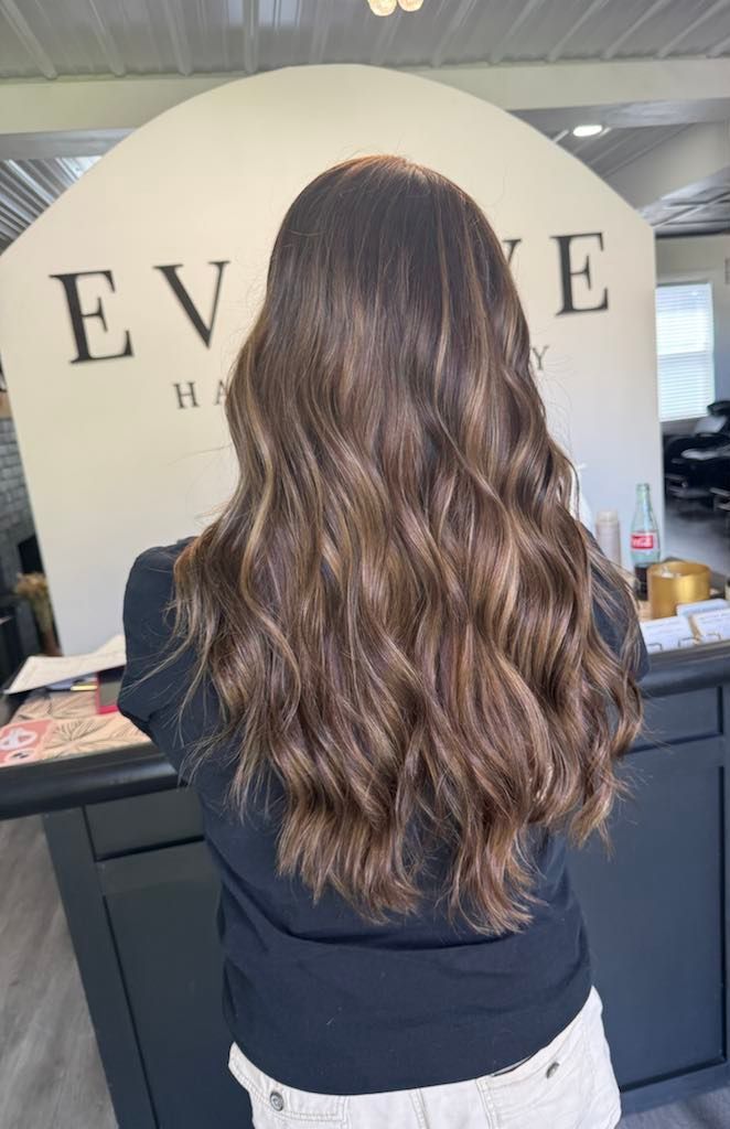 A person with long, wavy brown hair stands in front of a salon sign.