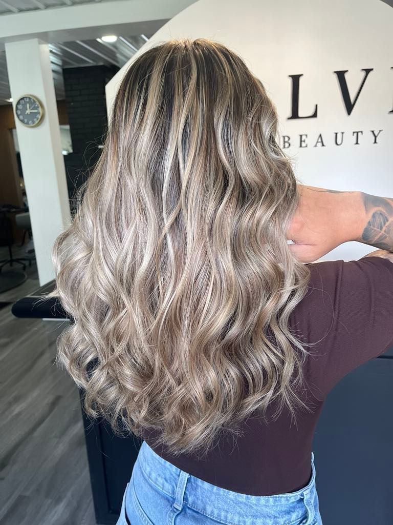 Woman with long wavy highlighted hair, mostly blonde and brown, indoors at a salon. She's wearing a brown top and blue jeans.