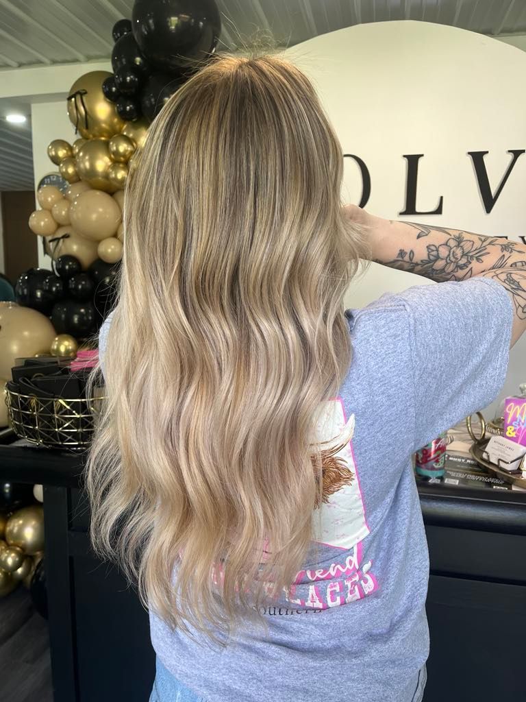Woman with long wavy blonde hair stands in front of a shop, wearing a gray t-shirt.